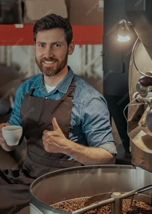 barista-drinking-coffee-sitting-near-coffee-roasting-machine-with-rise-finger-up_386167-18483